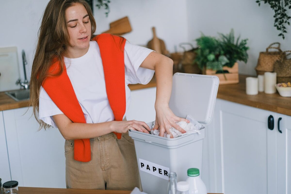 Young female in casual clothes sorting rubbish and putting paper into plastic bucket in light kitchen in daytime